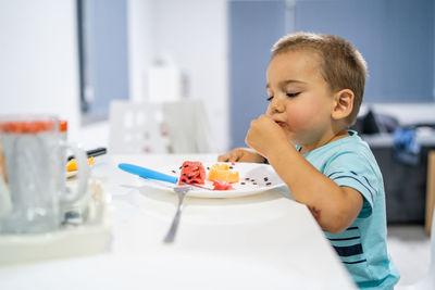 Side view of cute boy eating fruits in plate on table