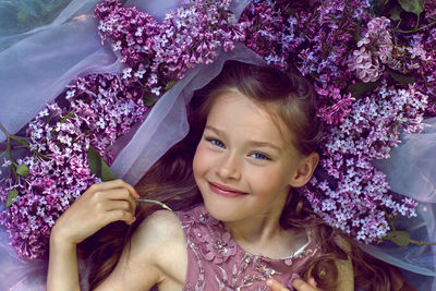Child girl in a purple floral dress lies on the ground among lilacs on a veil in spring
