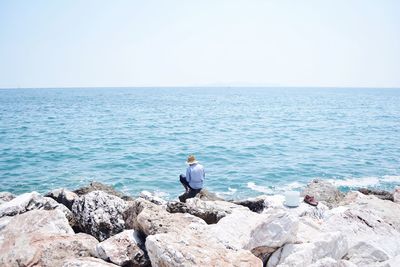 Rear view of man sitting at rocky beach against sky during sunny day