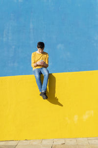 Full length of boy using phone while sitting against wall