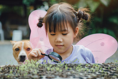 Cute girl holding magnifying glass by plants with dog in background