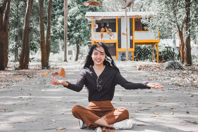 Portrait of smiling young woman throwing leaves while sitting on road in forest