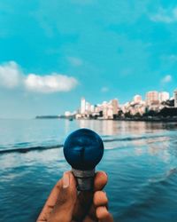 Cropped hand holding light bulb at sea against sky