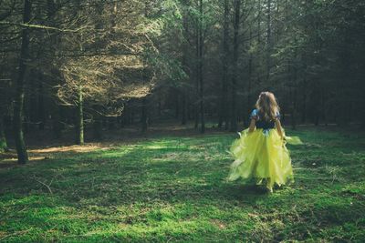 Rear view of girl in dress walking on grassy field at forest