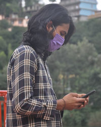 Side view of a long haired young man wearing face mask with using phone while standing outside 