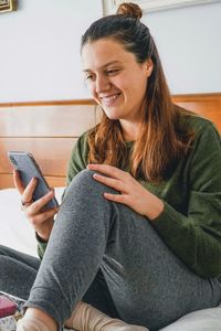 Smiling young woman using mobile phone sitting on bed at home