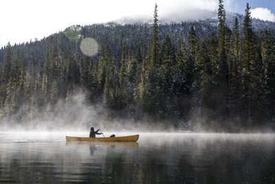 Man paddles canoe on lake with mist and fog on sunny day by forest