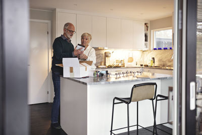 Full length of senior man by partner unpacking package over kitchen island