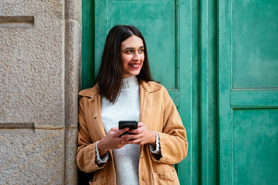 Young woman using mobile phone while standing against door