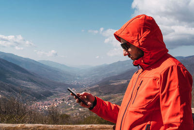 Caucasian male in a red coat typing with his smartphone with mountains in the background 