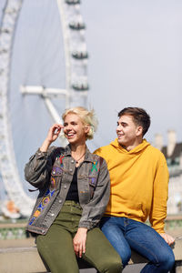 Smiling young couple sitting against ferris wheel in city