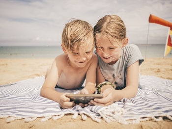 Smiling siblings sharing smart phone while lying on towel at beach against sky