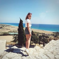 Side view of young woman standing at beach against clear sky during sunny day