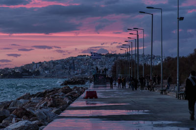 People on street by city against sky at sunset