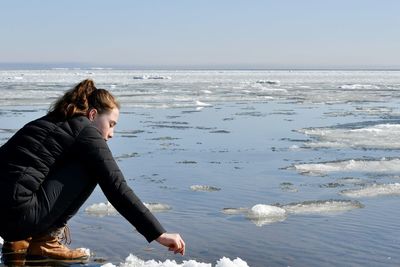 Full length of young woman sitting by frozen lake during winter