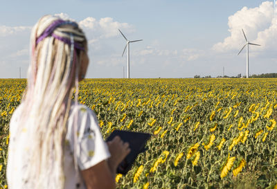 Young woman with tablet in field with sunflowers, wind turbines for green energy, eco-energy, space 