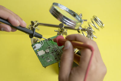 Cropped hands of person checking circuit board with work tool against yellow background