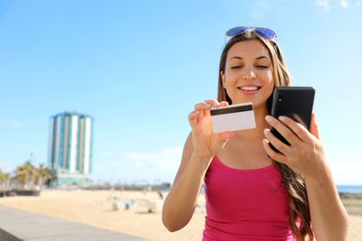 Beautiful young woman paying online with her telephone reading credit card number outdoor on summer.