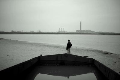 Rear view of man standing by water in shipwreck at beach against factory