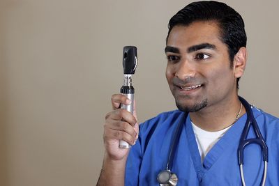 Smiling doctor looking through ophthalmoscope against beige background