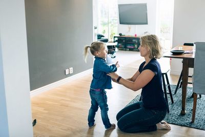 Side view of mother dressing daughter while kneeling on floor at home