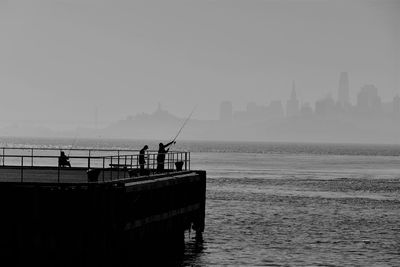 Silhouette people fishing on pier at sea against sky in city