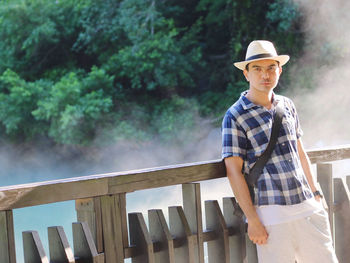 Portrait of young man standing by railing against trees