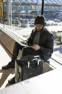 Stylish bearded guy in knitted hat and eyeglasses sending using phone while sitting with laptop against glass wall in airport