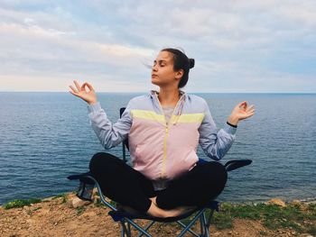 Young woman meditating while sitting on chair at beach against cloudy sky