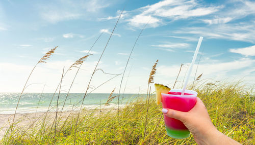 Hand holding a glass with cocktail and straw, sand dunes with grass on beach with ocean on