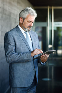 Mature businessman using digital tablet while standing against wall