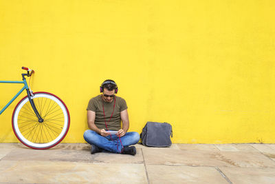 Man using phone while sitting with bicycle against yellow wall