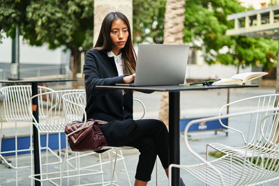 Serious hispanic woman in elegant wear working on modern netbook while sitting at table on terrace of cafe in city