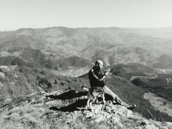 Side view of man sitting on cliff with dog against sky