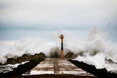 Waves splashing by lighthouse and pier against cloudy sky during sunset