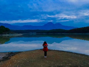 Rear view of woman standing at lakeshore against cloudy sky