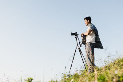 Surface level shot of man with tripod and camera standing on field against clear sky