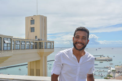 Portrait of smiling young man standing on pier against sky on sunny day