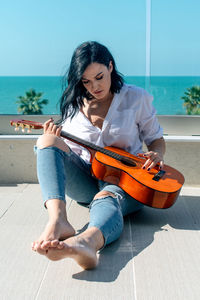 Beautiful woman with guitar sitting by railing against clear sky