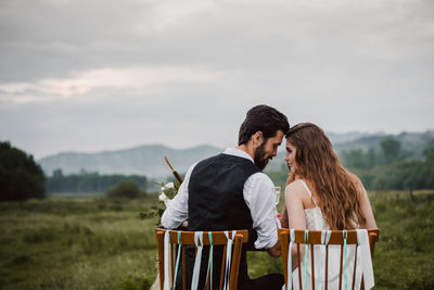 Rear view of bride and bridegroom sitting on chairs at field