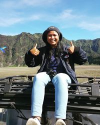 Portrait of smiling young woman showing thumbs up while sitting on car roof against sky