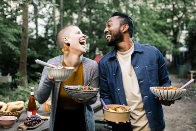 Multiracial male and female friends laughing and looking at each other while holding food bowls