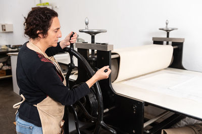 Side view of female artisan spinning wheel of printing press machine while making artwork in linocut technique in workshop