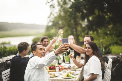 Man taking selfie with friends holding drinks during dinner party in backyard