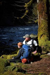 Father and son carrying backpacks resting on riverbank at forest during sunny day