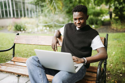 Young black man working on laptop outdoors, looking at screen, smiling