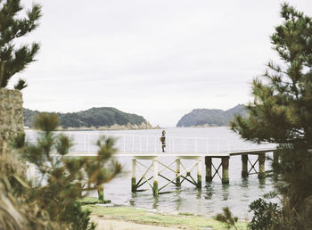 Mid distant view of woman standing on footbridge over river against sky