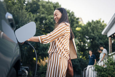 Low angle view of smiling woman charging car with electric plug in back yard