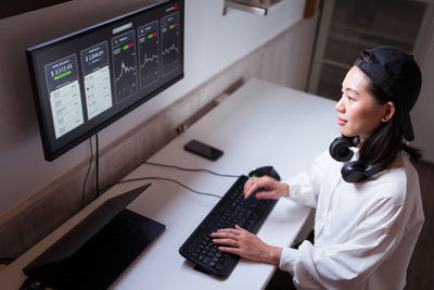 High angle concentrated asian female working on computer with charts showing dynamic of changes in value of cryptocurrency at convenient workplace