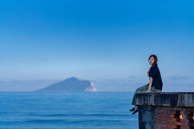 Side view portrait of young woman sitting on building terrace by sea against blue sky